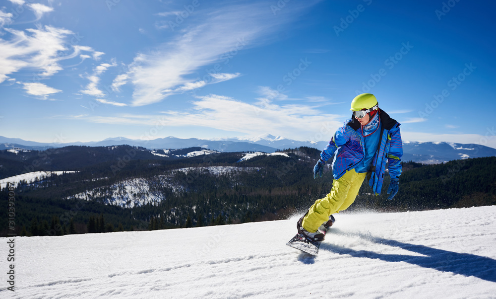 Snowboarder man in goggles, helmet and warm bright clothing riding snowboard on background of blue sky and snowy mountains covered with spruce trees on sunny winter day. Sport and recreation concept.