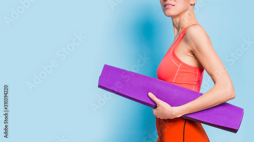  Portrait of happy fitness girl holding yoga exercising mat over light blue background wearing bright sporty outfit of orange color