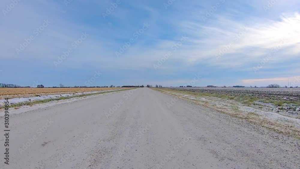 POV through the rear window while driving on a rural gravel road past empty fields on a sunny winter afternoon; there is a light dusting of snow in the ditches POV through the rear window while driving on a rural gravel road past empty fields on a sunny winter afternoon; there is a light dusting of snow in the ditches