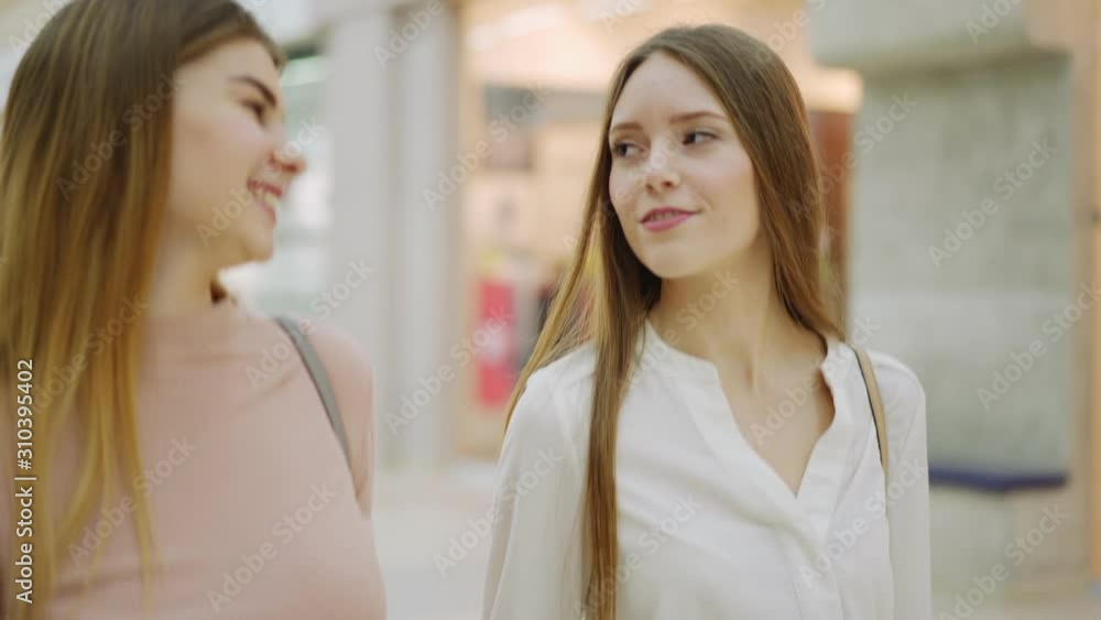 Tracking tilt up side view shot of two young women with shopping bags walking together in mall, talking joyfully and laughing Tracking tilt up side view shot of two young women with shopping bags walking together in mall, talking joyfully and laughing