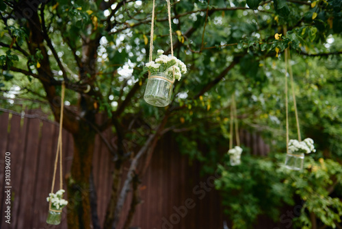 Rustic wedding decorations with flowers in glass jar hanging on green tree outdoors, copy space. Floral arrangement. Wedding concept