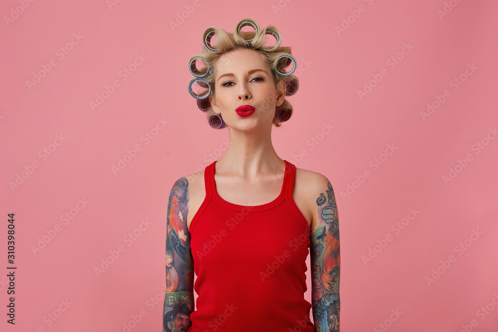 Studio shot of charming young blonde tattooed lady making hairdo and folding her red lips in air kiss while standing over pink background in red shirt with hands down Studio shot of charming young blonde tattooed lady making hairdo and folding her red lips in air kiss while standing over pink background in red shirt with hands down