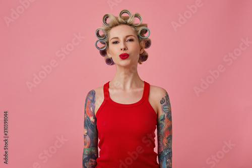 Studio shot of charming young blonde tattooed lady making hairdo and folding her red lips in air kiss while standing over pink background in red shirt with hands down