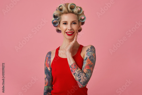 Charming young blonde tattooed woman with evening makeup making hairdo before upcoming party, touching soft her face and smiling gently at camera, isolated over pink background © timtimphoto