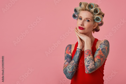Elegant young blonde female with tattooes folding hands under her chin and looking gently to camera  making hairdo and having festive makeup while standing over pink background