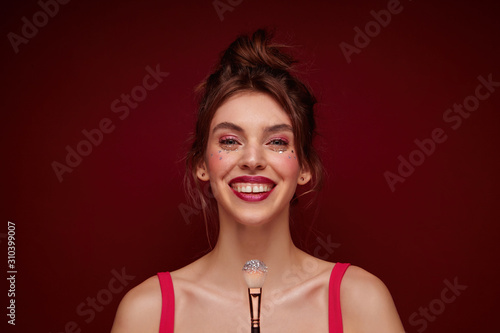 Close-up of positive attractive young brunette woman with bun hairstyle smiliing happily at camera and holding brush with lushes, making evening makeup while posing over burgundy background photo