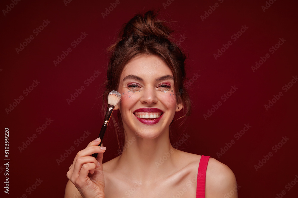 Happy young brown haired lovely woman with festive makeup wearing casual hairstyle while standing over burgundy background, meeting with friends in evening and being in high spirit Happy young brown haired lovely woman with festive makeup wearing casual hairstyle while standing over burgundy background, meeting with friends in evening and being in high spirit