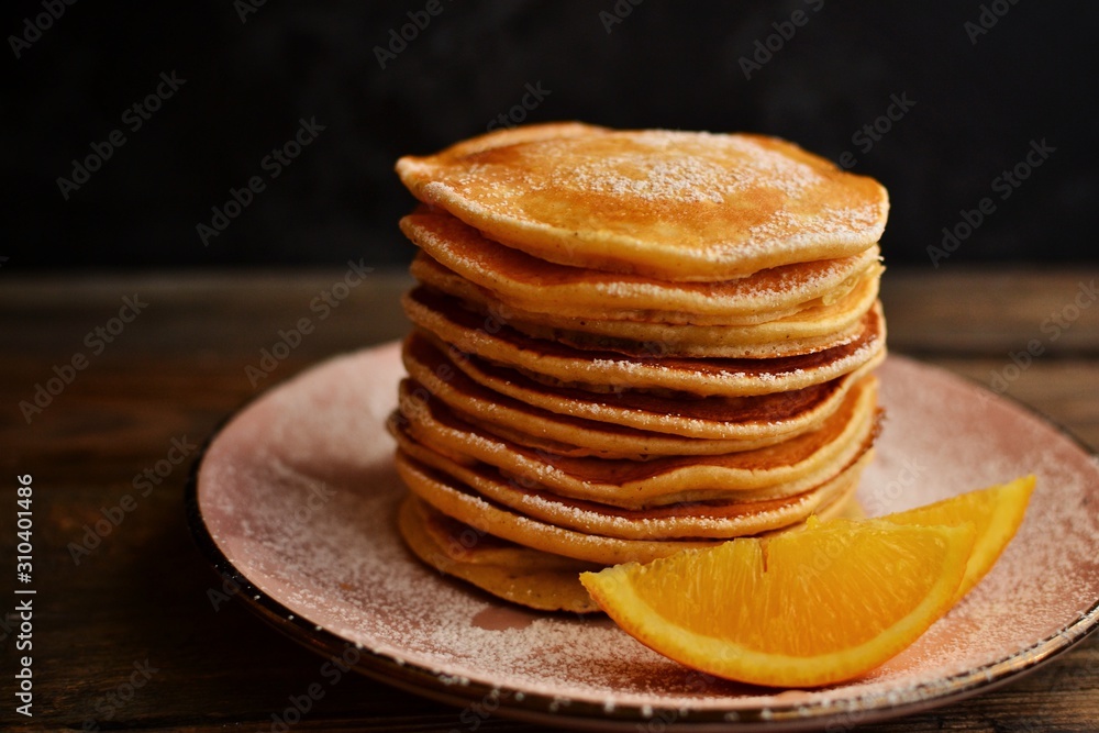 Pancakes with icing sugar and orange. Pancakes in a beautiful pink plate on a dark black wooden background. Delicious sweet food. Pancakes with icing sugar and orange. Pancakes in a beautiful pink plate on a dark black wooden background. Delicious sweet food.