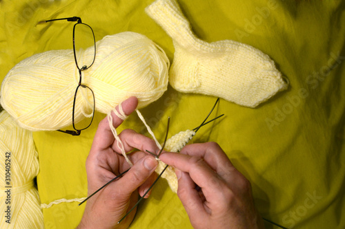 Knitting needles with knitting thread in mom's hands close-up on a mustard color background. Two skeins of thread of light yellow color, knitted sock and glasses closeup.