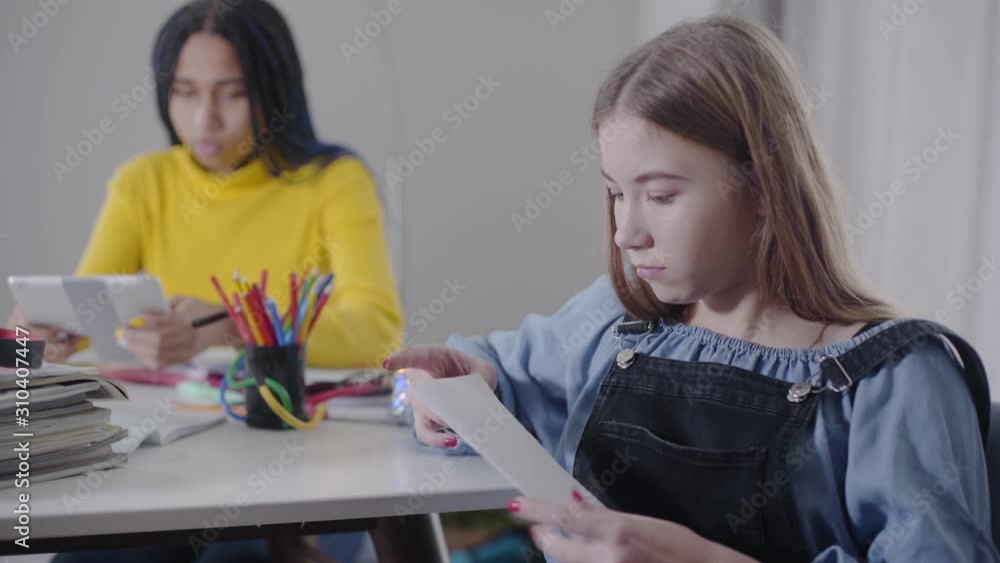 Portrait of surprised Caucasian girl opening sheet and looking away. Her African American friend sitting at the background and doing homework. Education, studying, learning. Portrait of surprised Caucasian girl opening sheet and looking away. Her African American friend sitting at the background and doing homework. Education, studying, learning.