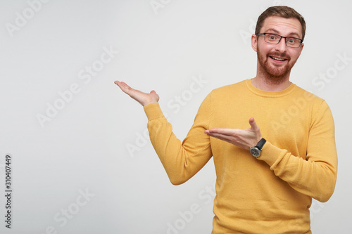 Studio shot of cheerful young handsome brunette male with beard showing aside with raised palms and looking happily at camera, dressed in mustard sweater and eyewear over white background