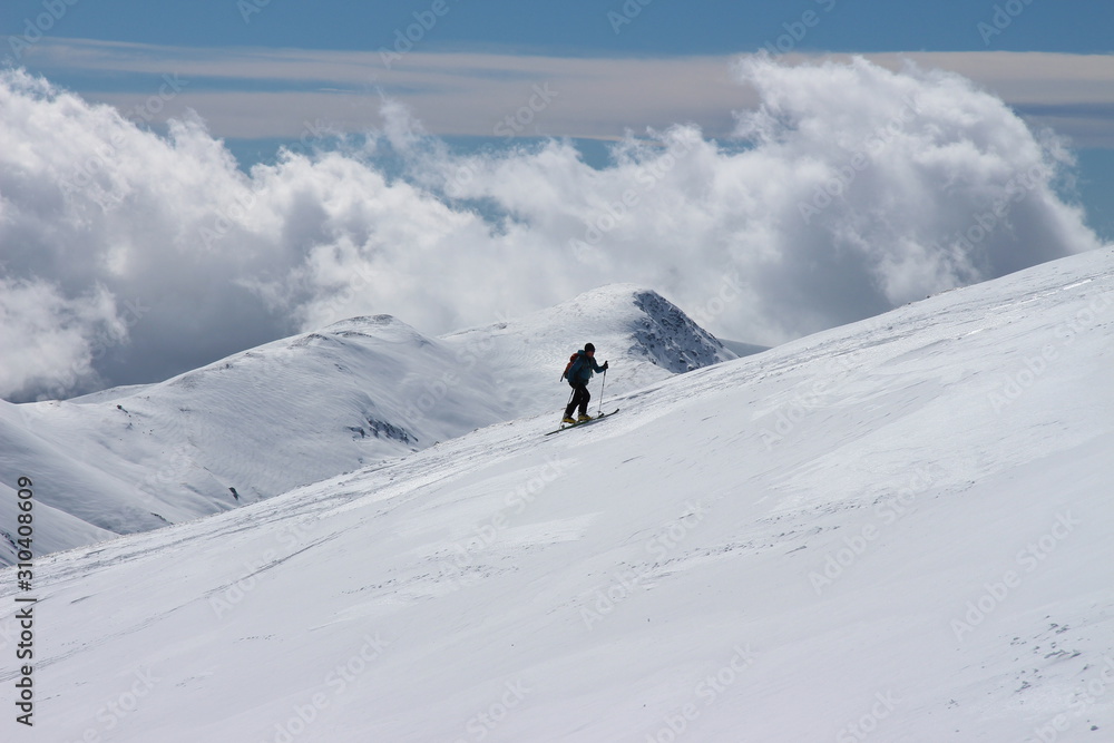 esquiador subiendo la ladera de una montaña nevada con nubes al fondo