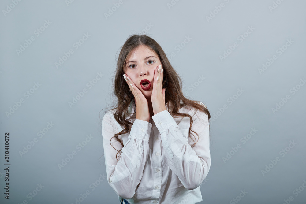 Shocked caucasian young woman in fashion white shirt holding hands on cheek with opened mouth, looking at camera isolated on gray background in studio. People sincere emotions, lifestyle concept. Shocked caucasian young woman in fashion white shirt holding hands on cheek with opened mouth, looking at camera isolated on gray background in studio. People sincere emotions, lifestyle concept.