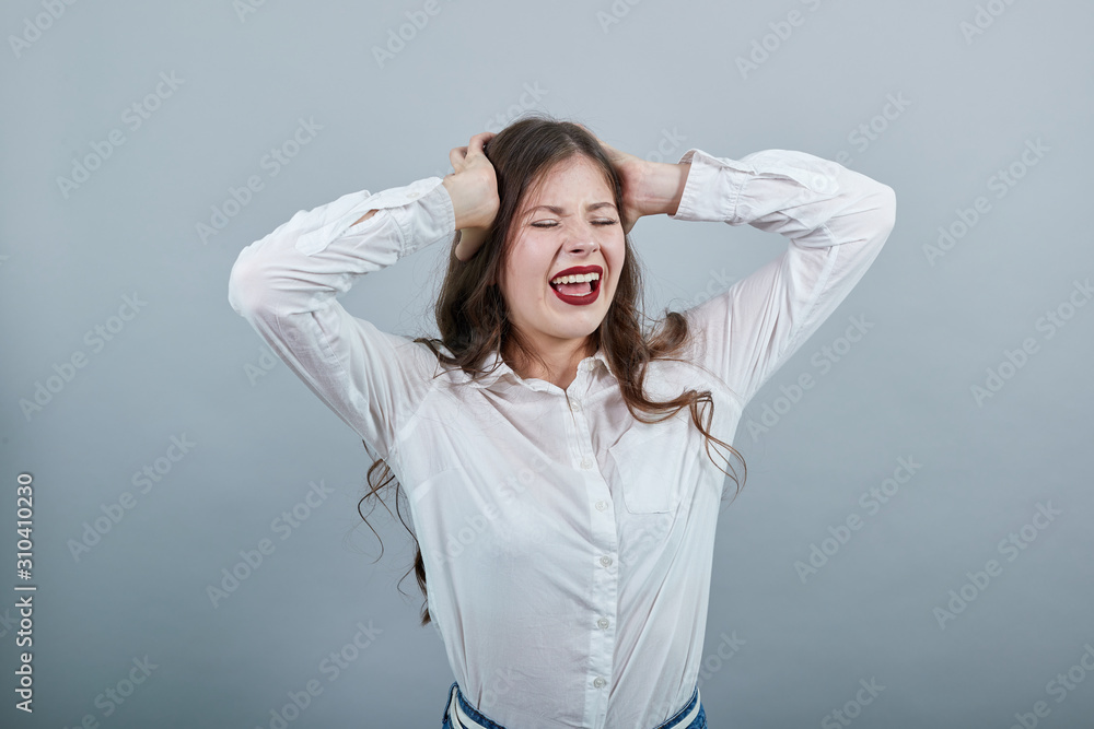 Charming caucasian young woman in fashion white shirt isolated on gray background in studio holding hands on head, screaming with opened mouth. People sincere emotions, lifestyle concept. Charming caucasian young woman in fashion white shirt isolated on gray background in studio holding hands on head, screaming with opened mouth. People sincere emotions, lifestyle concept.