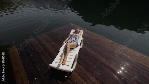 Woman lie on a sunbed in sunglasses and a boho silk shawl. Girl rest on a flood wood underwater pier. The pavement is covered with water in the lake. In the background are mountain and a forest. photo