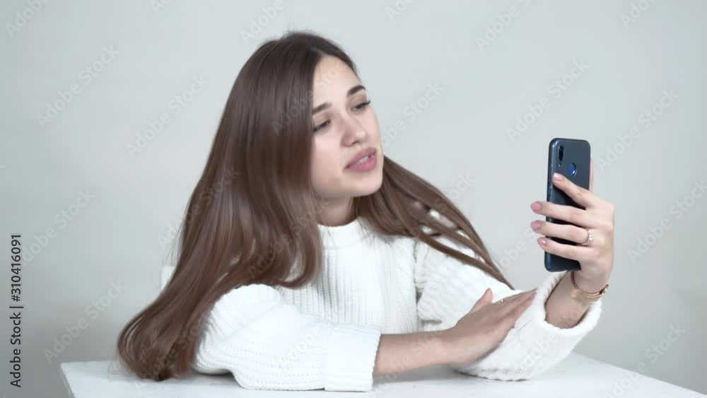 Caucasian girl white jumper. Her hair is straight and falls over her shoulders.She sits at a table, holds a smartphone, looks at the camera, gestures emotionally. Close up. Grey background