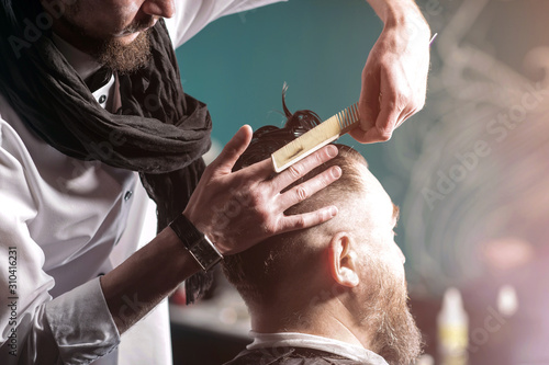 Barbershop. Man hairdresser in a white shirt and black scarf doing hairstyle a man with a beard.