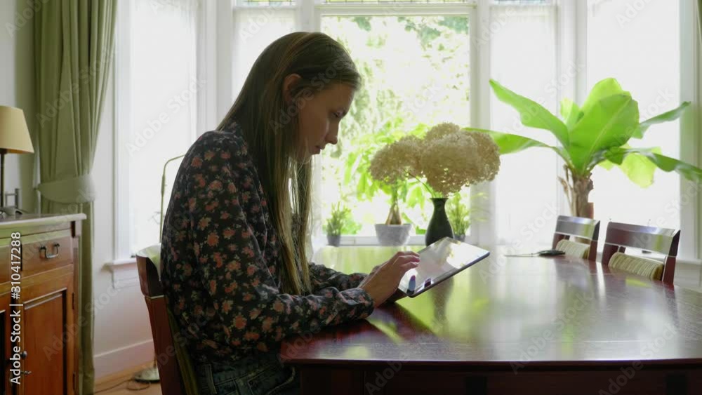 Young attractive caucasian woman using a tablet while sitting at a wooden table in a room with plants in slow motion Young attractive caucasian woman using a tablet while sitting at a wooden table in a room with plants in slow motion
