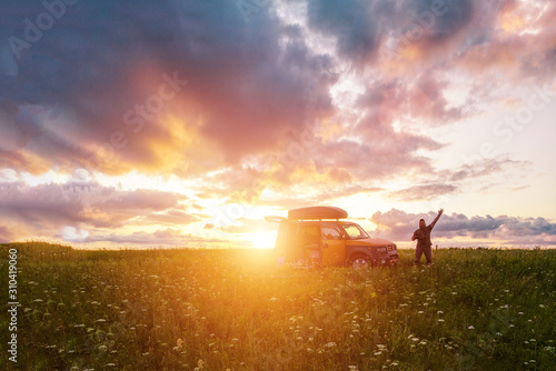 Man in a field with a raised hand near a car at sunset.