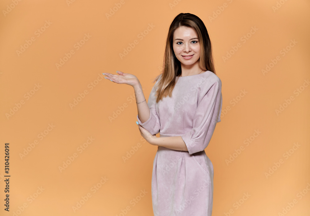 Portrait of a pretty young smiling woman on a beige background in a pink dress with long straight hair. Standing right in front of the camera, Shows emotions, talks in different poses.