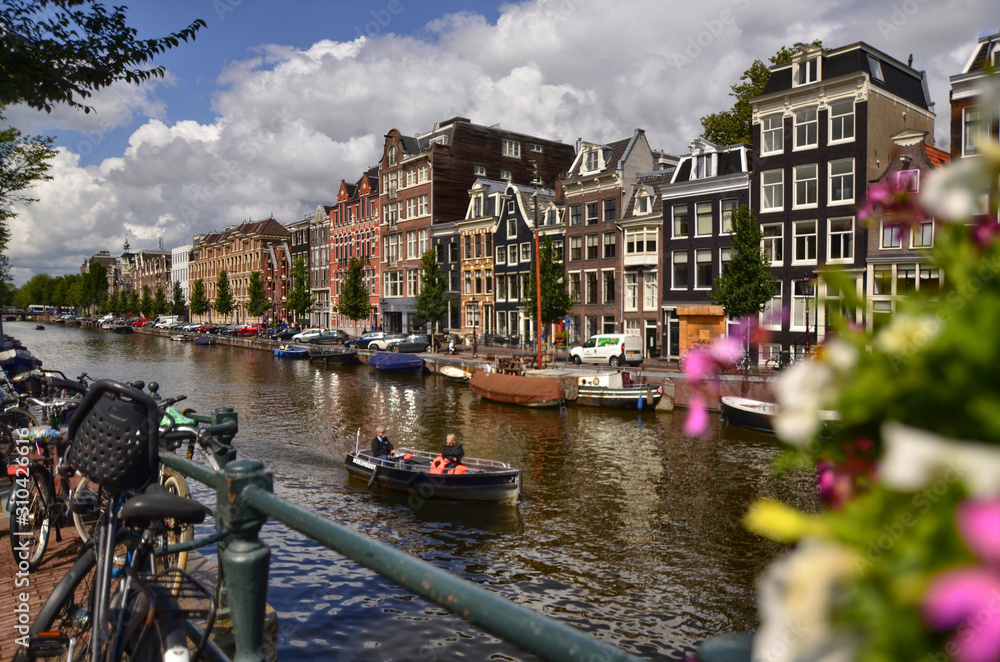 Fototapeta premium Amsterdam,Holland,August 2019. Enchanting view of a canals of the historic center. The typical houses overlook the water, boats moored along the canal. Planters on railings and bikes. Sunny day.