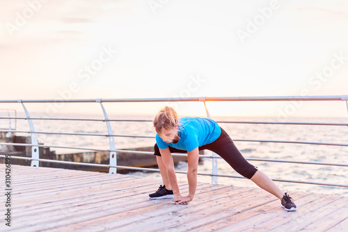 Young girl with long blonde hair in blue shirt and black leggins doing stretching exercise deep side lunge withforward bend on seashore at sunrise