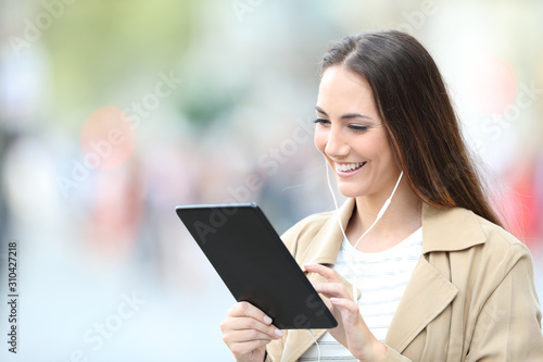 Happy woman wearing earbuds using tablet in the street