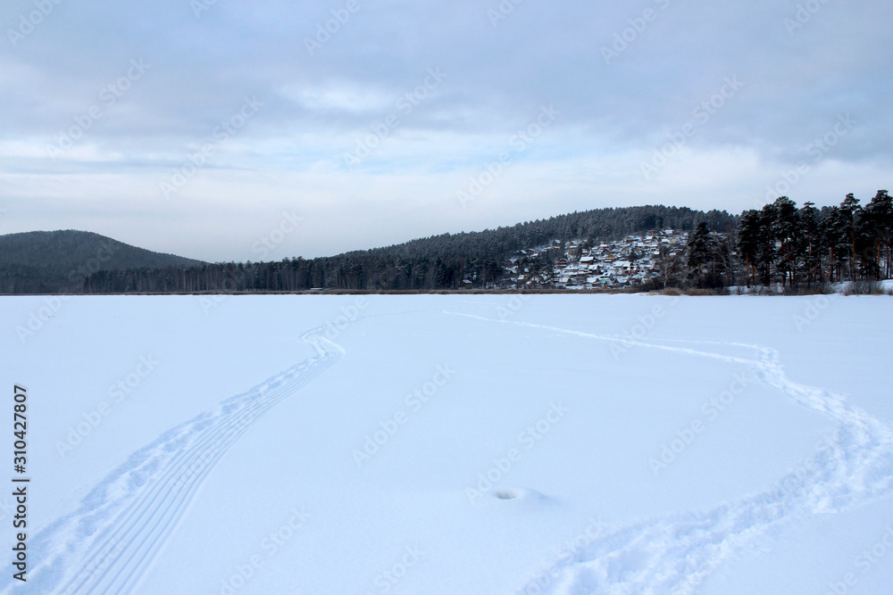 Winter landscape on the lake, forest and country houses on the hills in the background. Snow trail and trails extending into the distance to small houses. White silence, snow and sky