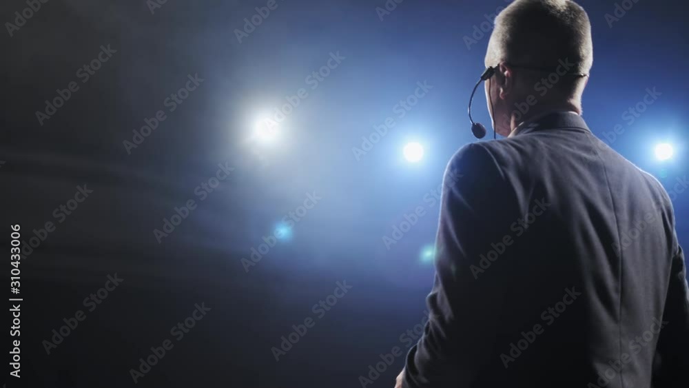 Rear view of men speaking through a microphone in dark conference hall. Man talks into microphones at press conference. Profile of mature male politician speaking passionately from tribune Rear view of men speaking through a microphone in dark conference hall. Man talks into microphones at press conference. Profile of mature male politician speaking passionately from tribune