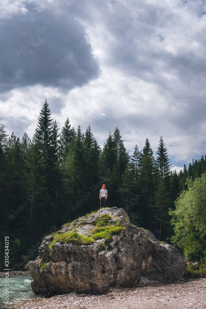 Girl on a large stone, a boulder, among the incredibly beautiful river of turquoise color, pine forest. summer day. vertical photo. Dolomites mountains, Camping Olympia Fiames, Cortina Ampezzo Italy.