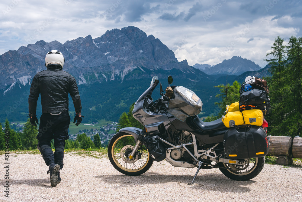 Back view of biker with adventure touring motorcycle in full equipment on dirt road, High top mountains on background, tourism travel concept, copy space. world riders