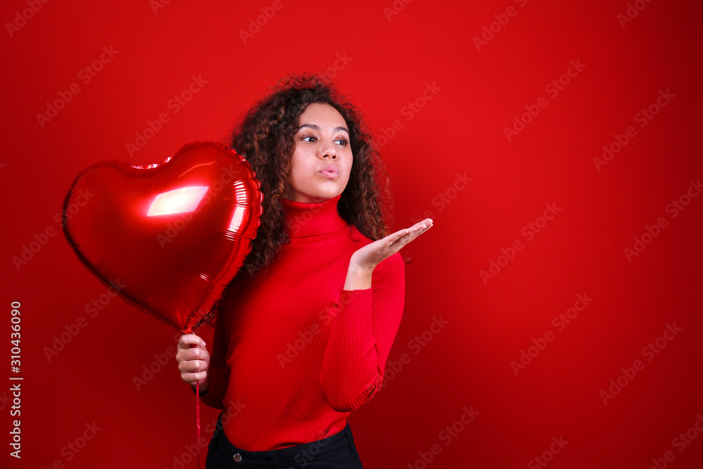 Studio portrait of young woman with dark skin and long curly hair wearing knitted turtle neck sweater over the festive red wall with heart shaped balloon. Close up, isolated background, copy space. Studio portrait of young woman with dark skin and long curly hair wearing knitted turtle neck sweater over the festive red wall with heart shaped balloon. Close up, isolated background, copy space.