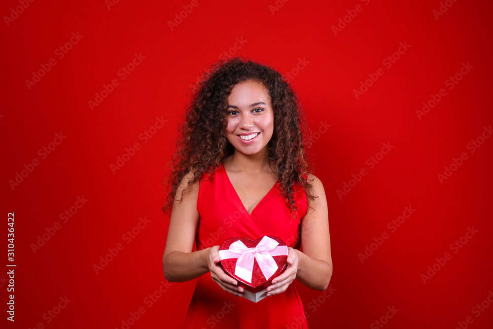 Studio portrait of young woman with dark skin and long curly hair wearing sexy dress over the festive red wall holding heart shaped candy box. Close up, isolated background, copy space. Studio portrait of young woman with dark skin and long curly hair wearing sexy dress over the festive red wall holding heart shaped candy box. Close up, isolated background, copy space.