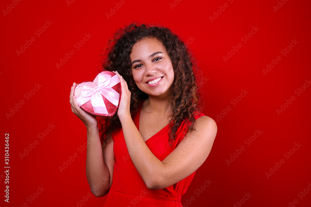 Studio portrait of young woman with dark skin and long curly hair wearing sexy dress over the festive red wall holding heart shaped candy box. Close up, isolated background, copy space. Studio portrait of young woman with dark skin and long curly hair wearing sexy dress over the festive red wall holding heart shaped candy box. Close up, isolated background, copy space.