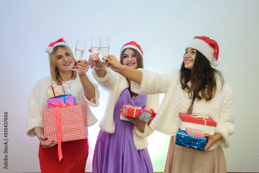 Obraz premium Three women in elegant dresses and santa hats with glasses of champagne. Merry Christmas concept.