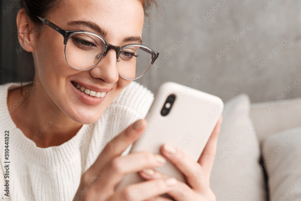 Image of woman in eyeglasses smiling and holding cellphone at home Image of woman in eyeglasses smiling and holding cellphone at home