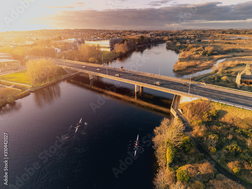 Bridge in Galway city over river Corrib at sunset. Cloudy sky. Rowing team training. Warm tones. Sun flare. Aerial view.