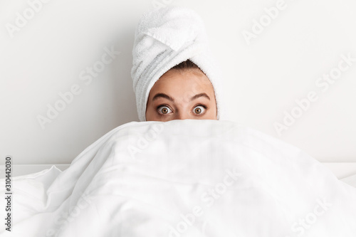 Image of surprised young woman hiding under blanket in bed at home