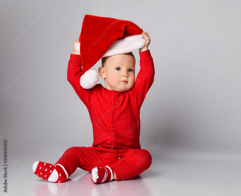 Little boy toddler in red overall and warm christmas socks is putting on playing taking off santa claus cap with pompon Little boy toddler in red overall and warm christmas socks is putting on playing taking off santa claus cap with pompon
