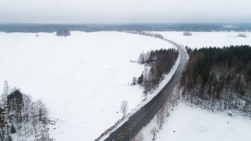 Aerial view of curving road, forests and lakes. Road on a cloudy day. Beautiful winter landscape terrain with drone.