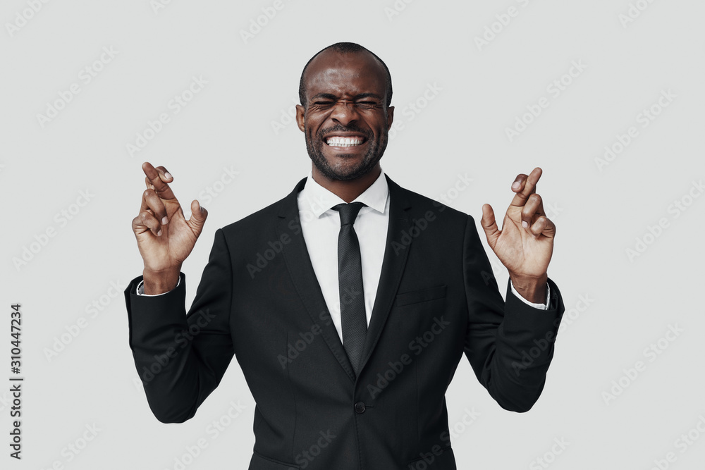 Handsome young African man in formalwear making a face and keeping fingers crossed while standing against grey background Handsome young African man in formalwear making a face and keeping fingers crossed while standing against grey background