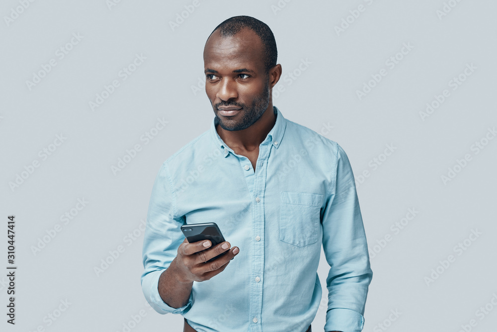 Handsome young African man using smart phone while standing against grey background Handsome young African man using smart phone while standing against grey background