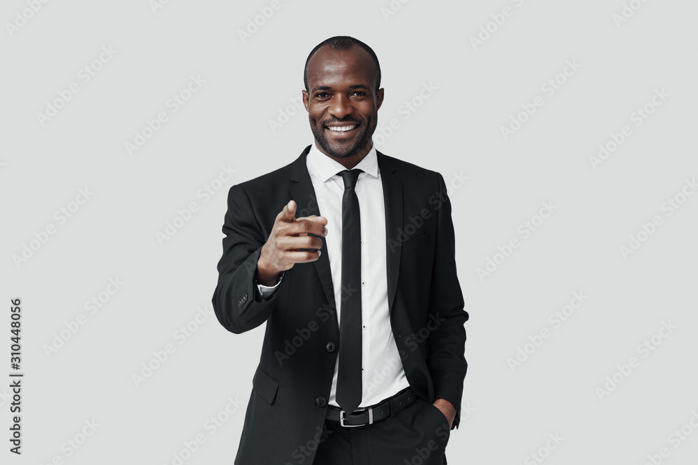 Handsome young African man in formalwear pointing you and smiling while standing against grey background Handsome young African man in formalwear pointing you and smiling while standing against grey background