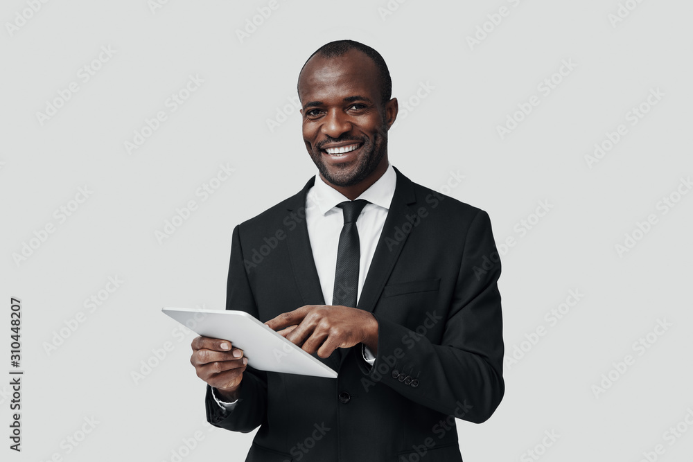 Charming young African man in formalwear working using digital tablet while standing against grey background Charming young African man in formalwear working using digital tablet while standing against grey background