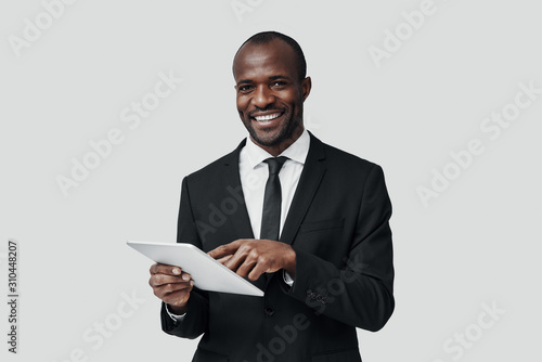 Charming young African man in formalwear working using digital tablet while standing against grey background