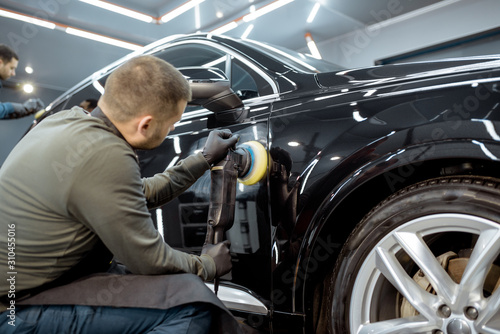 Two workers polishing vehicle body with special grinder and wax from scratches at the car service station. Professional car detailing and maintenance concept