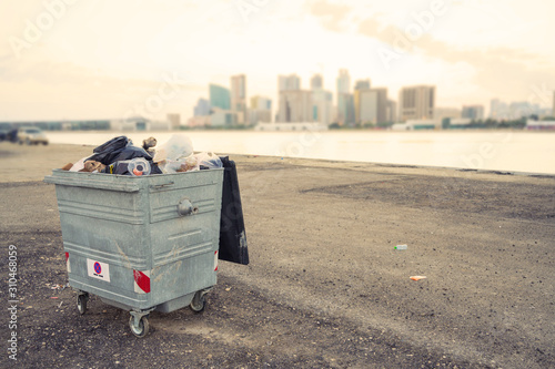 Big metal bin full of rubbish outside park with city in background over sky, Bahrain.