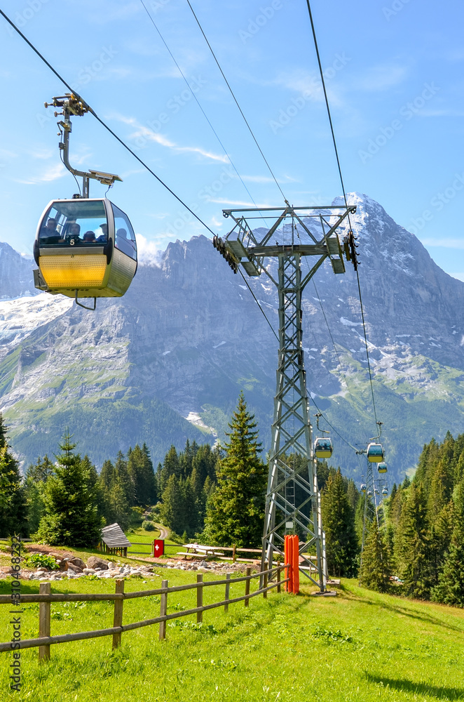 Yellow cable car in the Swiss Alps. Gondola going from Grindelwald to First in the Jungfrau region. Summer Alpine landscape with snowcapped mountains in the background. Transportation of tourists