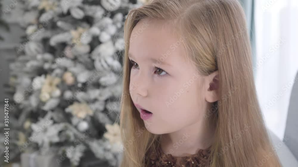 Camera moving around face of cute Caucasian girl with long hair. Pretty child sitting next to the Christmas tree on New Year's eve. Beauty, holidays season. Camera moving around face of cute Caucasian girl with long hair. Pretty child sitting next to the Christmas tree on New Year's eve. Beauty, holidays season.