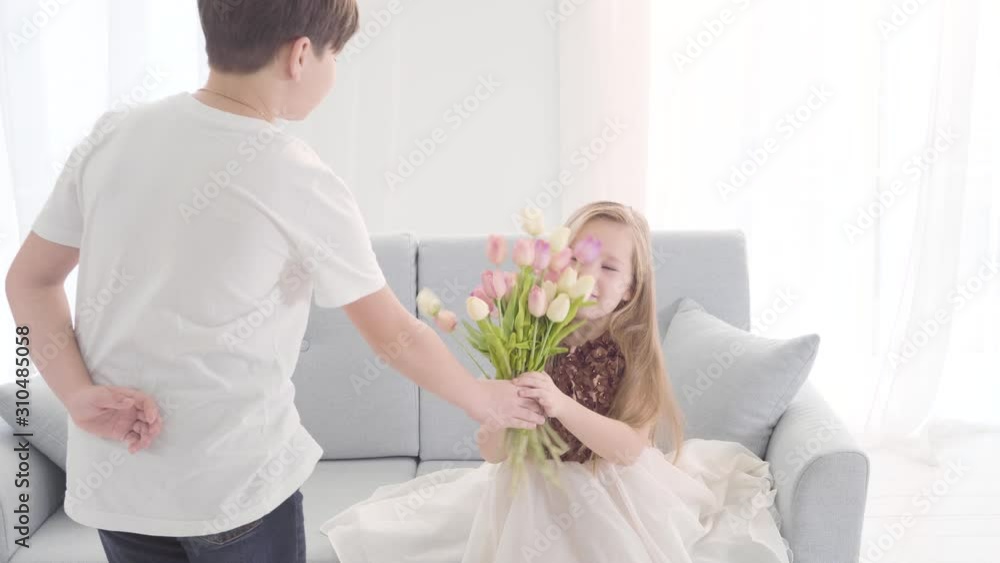 Caucasian boy giving bouquet of tulips to charming girl sitting on sofa. Little lady taking gift and smelling flowers. Flirting, first love, innocence, childhood. Caucasian boy giving bouquet of tulips to charming girl sitting on sofa. Little lady taking gift and smelling flowers. Flirting, first love, innocence, childhood.